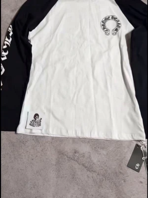 Chrome Hearts långärmad t-shirt - Snygg långärmad t-shirt från Chrome Hearts med svartvita kontrastärmar och stora tryck på både bröstet och ryggen. Ärmarna har cool text längs sidorna och tröjan har en relaxed fit. Perfekt för dig som gillar streetwear och vill sticka ut.