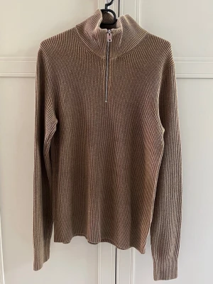 Brun ribbstickad tröja med halvzip - Säljer en beige ribbstickad tröja med halv dragkedja och hög krage. Tröjan har lång ärm och är tillverkad i ett mjukt, stretchigt material som ger en clean och stilren look. 