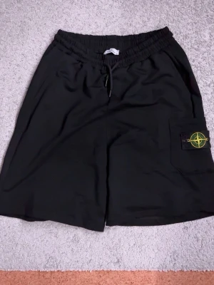 Svarta shorts från Stone Island - Säljer ett par svarta shorts från Stone Island med elastisk midja och snörning. Shortsen har en ficka på sidan med den klassiska Stone Island-loggan i gult och grönt. Perfekta för sommaren och riktigt snygga till streetwear-stilen.
