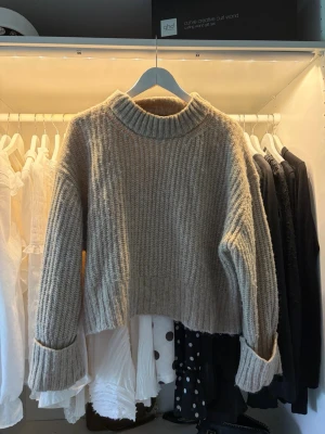 Beige ribbstickad tröja med polokrage - Mysig beige stickad tröja med bred ribbstickning och hög polokrage. Tröjan har långa ärmar med uppvikta muddar och en något boxig, croppad passform. Perfekt för lager på lager och enkel att styla till olika outfits. Storlek L men som en S