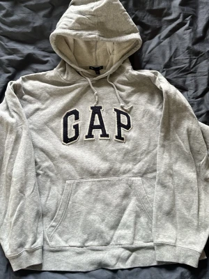 Grå hoodie från GAP med huva - Säljer en klassisk grå hoodie från GAP med stor logga i svart och vitt på bröstet. Hoodien har huva med dragsko, magficka och ribbade muddar vid ärmar och nederkant. 