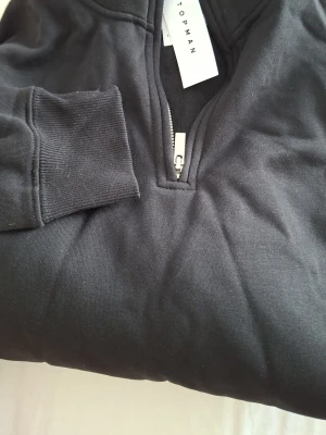 Svart hoodie med dragkedja från Topman - Snygg svart hoodie från Topman i storlek M. Hoodien har en halv dragkedja framtill, ribbade muddar vid ärmsluten och är gjord i mjukt bomullsmaterial. Perfekt för en avslappnad och stilren look.