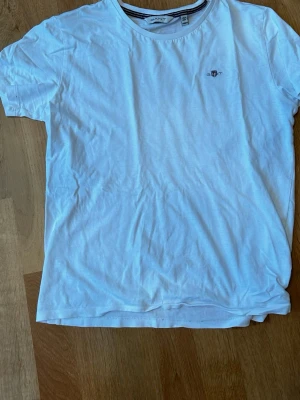 Vit t-shirt från Gant - Klassisk vit t-shirt från Gant med rund halsringning och diskret logga broderad på bröstet. Tillverkad i mjuk bomull och har en normal passform med korta ärmar. Perfekt basplagg till garderoben. Hör av er vid funderingar 😁