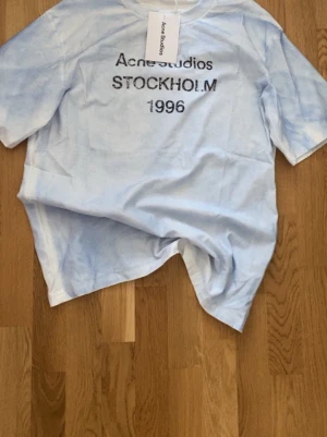 Ljusblå Acne Studios t-shirt - Säljer en ljusblå t-shirt från Acne Studios med tryck 'STOCKHOLM 1996' på bröstet. T-shirten har en avslappnad passform och är tillverkad i mjuk bomull. Perfekt för en clean och stilren look. Passar både tjejer och killar och är allmänt snygg 🤩 / HELT NY!! (Äkta såklart) 