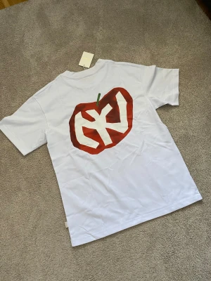 Vit Aime Leon Dore t-shirt med grafiskt äppletryck - Vit t-shirt med ett stort grafiskt äppletryck i rött och grönt på ryggen och texten '214 MULBERRY ST. NY' på bröstet. Klassisk passform och rund halsringning. Perfekt för dig som gillar unika prints och streetstyle.