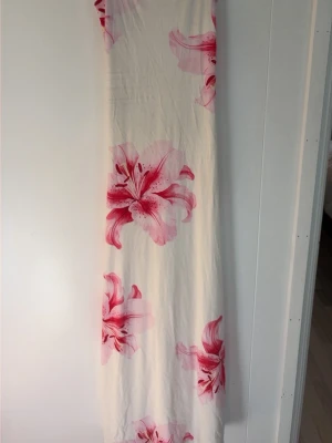 Vit långklänning med rosa blommor - Säljer en elegant vit långklänning med stora rosa blommor. Klänningen har en rak och enkel passform och är ärmlös, perfekt för varma dagar. Materialet känns mjukt och svalt, troligen bomull eller polyester. Perfekt för sommarens alla tillfällen! 