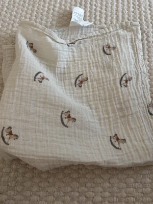 Beige muslinduk med gunghästar - Säljer en beige muslinduk med gulligt mönster av små gunghästar i brunt och grått. Tyget är mjukt och luftigt, perfekt som accessoar eller filt. Passar till dig som gillar söta detaljer och stilren design.