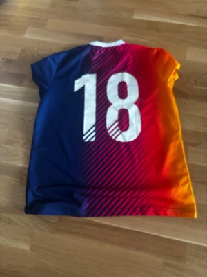 EA Sports FIFA 18 tröja i regnbågsfärg - Säljer en snygg FIFA 18 tröja från EA Sports med coolt färgskifte från blått till rött och orange. Stora vita siffror '18' på ryggen och EA Sports-logga framtill. Tillverkad i lätt och snabbtorkande material, perfekt för sport eller gaming.