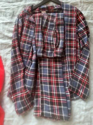 Rutig flanellpyjamas från Kappahl - Klassisk tvådelad pyjamas från Kappahl i mjuk flanell. Setet har en långärmad skjorta med knappar och matchande byxor med resår i midjan. Mönstret är rutigt i marinblått, rött och vitt. Perfekt för mysiga kvällar hemma.