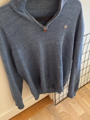 Blå stickad tröja med halvzip - Mysig blå stickad tröja med halvzip och hög krage. Tröjan har långa ärmar, ribbade muddar och en liten broderad detalj på bröstet. Perfekt för lager på lager och enkel att matcha med jeans eller chinos.