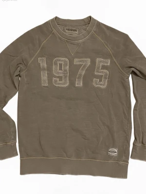 Olivgrön sweatshirt med 1975-tryck - Cool olivgrön sweatshirt med stort '1975' tryck på bröstet. Tröjan har rund halsringning, raglanärmar och ribbade muddar vid ärmslut och nederkant. Tillverkad i mjukt bomullsmaterial och har en avslappnad passform.