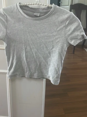 Ljusgrå ribbad t-shirt - En enkel och stilren ljusgrå t-shirt med ribbad struktur och rund hals. T-shirten har korta ärmar och är tillverkad i mjukt bomullsmaterial. Perfekt till jeans eller kjol för en avslappnad look.