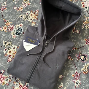 Mörkgrå hoodie från Polo Ralph Lauren - Snygg mörkgrå hoodie från Polo Ralph Lauren med dragkedja och huva. Klassisk broderad logga på bröstet, två fickor framtill och ribbade muddar. Tillverkad i mjuk bomull, perfekt för chill dagar eller när du vill ha en clean look.