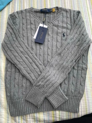 Grå kabelstickad tröja Polo Ralph Lauren - Snygg grå kabelstickad tröja från Polo Ralph Lauren med V-ringning och klassisk marinblå logga broderad på bröstet. Tröjan har ribbade muddar vid ärmslut och nederkant och är tillverkad i mjukt material som passar perfekt till många outfits.