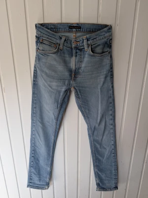 Ljusblå skinny jeans från Nudie Jeans - Säljer ett par ljusblå skinny jeans från Nudie Jeans med klassisk femficksmodell och orangea kontrastsömmar. Jeansen har en något tvättad look och är tillverkade i mjukt denimtyg. Perfekta för en avslappnad och trendig stil.