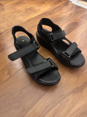 Svarta platåsandaler med kardborre - Säljer ett par svarta sandaler med platåsula och justerbara kardborreband. Sandalerna har öppen tå och häl, samt detaljer i metall vid banden. Perfekta för dig som gillar en sportig och bekväm stil.