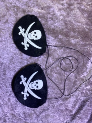 Pirat ögonmask  med dödskallemotiv - Svarta ögonmask i filtmaterial med klassiskt piratmotiv – vit dödskalle och korsade sablar. Perfekt för maskerad eller cosplay med pirattema. Fästs med tunna svarta snören. Finns fler kvar, samma frakt, 39kr/2st 