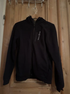 Svart hoodie med dragkedja - Svart hoodie med hel dragkedja framtill och huva. Har en bröstficka med dragkedja och tryckt text på bröstet. Perfekt för en avslappnad och sportig stil. Mjuk och bekväm passform.