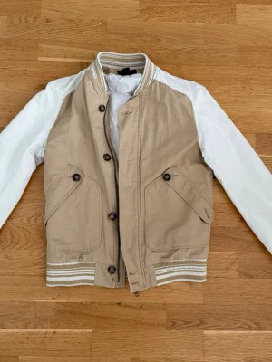Beige jacka från H&M - Snygg beige jacka från H&M med vita ärmar och ribbade muddar i beige och vitt. Jackan har två stora fickor framtill med knappstängning och stängs med både dragkedja och knappar. OBS: Det finns lite trådar lösa vid knapparna men stör inte looken. Perfekt för dig som vill ha en stilren och sportig look.
