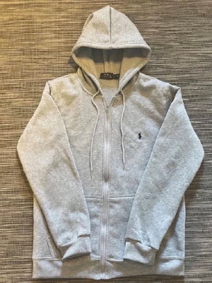 Grå zip hoodie från Polo Ralph Lauren - Snygg gråmelerad hoodie med dragkedja från Polo Ralph Lauren. Klassisk design med huva och dragsnören, två fickor framtill och den ikoniska lilla loggan broderad på bröstet. Perfekt för en chill och avslappnad stil.