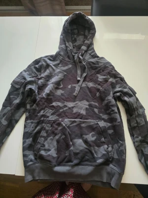 Camo hoodie från Peak Performance - Snygg hoodie från Peak Performance i grått camomönster. Tröjan har huva med dragsko och en stor magficka framtill. Hittat ett minimalt hål bakpå luvan (se bild)