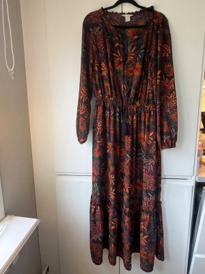 Långklänning med blommigt mönster - Långklänning med färgstarkt blommigt mönster i färger perfekt när hösten kryper på. Klänningen har lång ärm, v-ringning med knytband och resår i midjan för en bekväm passform. 