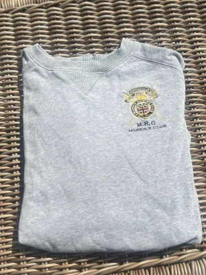 Ljusgrå sweatshirt Morris R Club - Ljusgrå sweatshirt med broderad Morris R Club-logga på bröstet och rund halsringning. Tröjan har ribbade muddar vid ärmslut och nederkant samt en liten patch nertill. Perfekt för en avslappnad och stilren look.
