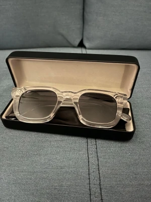 Transparenta solglasögon All Cap One - Snygga solglasögon från All Cap One med transparent, fyrkantig båge och mörka glas. Kommer i ett svart fodral med vit logga. Perfekta för dig som vill ha en modern och stilren look.