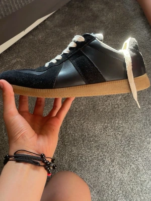 Maisson Margiella Gats mmg  - Snygga svarta sneakers med vit sula och vita skosnören. Ovandelen är i en mix av mocka och slätt läder, med klassisk gummisula i beige. Perfekta för dig som gillar stilrena och tidlösa skor med en sportig vibe. Pris går alltid i diskutera vid snabb affär☺️