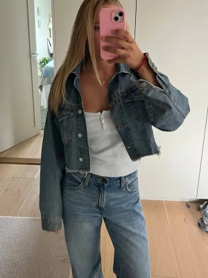 Cropped blå jeansjacka - Säljer en trendig cropped jeansjacka i klassisk blå denim. Jackan har rå kant nertill, silverfärgade knappar och två bröstfickor med lock. Perfekt oversized passform och cool vintagekänsla. Passar till allt och ger en avslappnad look.