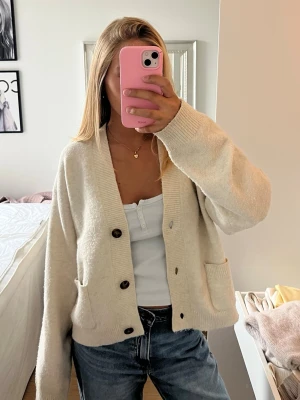 Beige kofta med fickor - Mysig beige oversized kofta med stora bruna knappar och två framfickor. Koftan har ribbade muddar och en djup v-ringning. Perfekt att slänga över en topp för en avslappnad look. Passar till jeans eller kjol.