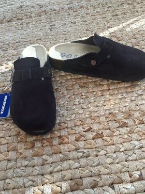 Svarta Birkenstock tofflor i mocka - Säljer ett par svarta Birkenstock tofflor i mjuk mocka med justerbart spänne och ergonomisk fotbädd. Klassisk slip-in modell som är både stilren och bekväm. Perfekta för dig som gillar en clean look och vill ha något som funkar till det mesta.