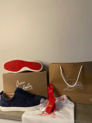 Blåa Christian Louboutin sneakers - Snygga blåa sneakers från Christian Louboutin i mocka med klassiska röda sulor och coola nitar framtill. Skorna har vita sulor och lågt skaft. Perfekta för dig som vill sticka ut med exklusiv design och detaljer.