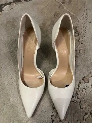 Vita spetsiga pumps från MNG - Säljer ett par stilrena vita pumps från MNG med spetsig tå och hög klack. Skorna har öppen insida och är tillverkade i ett slätt, glansigt material som ger en elegant look. Perfekta för dig som vill ha en klassisk men trendig sko.