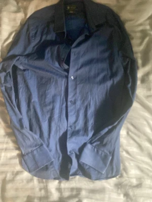 Blå skjorta från Riley Slim Fit - Säljer en stilren blå skjorta från Riley i modellen Slim Fit. Skjortan har långa ärmar, klassisk krage och knäppning framtill. Perfekt för dig som gillar en smalare passform och enkel, snygg stil.