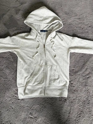 Grå hoodie från Polo Ralph Lauren - Snygg ljusgrå hoodie från Polo Ralph Lauren med dragkedja, huva och dragsnören. Klassisk broderad logga på bröstet och känguruficka framtill. Perfekt för en chill och stilren look. Mjuk bomullsblandning som känns skön mot huden.