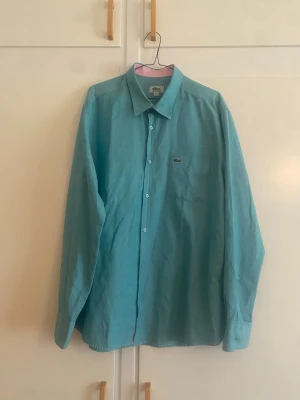 Lacoste Ljusblå skjorta med knappstängning - Säljer en    Lacoste ljusblå skjorta med klassisk krage och knappstängning framtill. Skjortan har en bröstficka med liten broderad logga och långa ärmar med manschetter. Perfekt för en clean och stilren look.