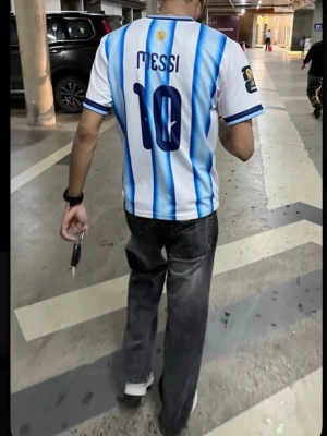 Argentina Messi fotbollströja - Säljer en klassisk Argentina fotbollströja med blå och vita ränder, nummer 10 och Messi på ryggen. Tröjan har korta ärmar och rund hals, samt detaljer i blått vid ärmslut och krage. Perfekt för dig som älskar fotboll och vill stötta ditt favoritlag.