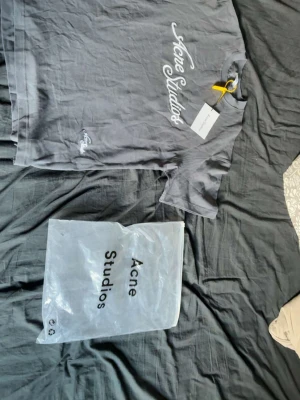 Acne Studios  - Snygg grå t-shirt från Acne Studios med vit broderad logga på bröstet. Klassisk passform och korta ärmar. Tillverkad i mjuk bomull för en skön känsla. Perfekt för dig som gillar stilrena och trendiga plagg.