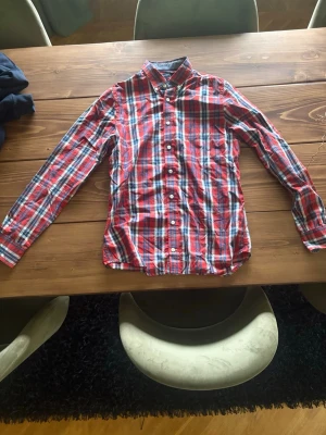 Rutig röd skjorta Tommy Hilfiger - Säljer en rutig skjorta från Tommy Hilfiger i rött, blått och vitt. Skjortan har lång ärm, klassisk krage och knappar framtill. Slim fit-modell i mjuk bomull, perfekt för en snygg och avslappnad stil.