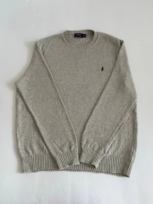 Stick Ralph Lauren tröja - Ljusgrå stickad tröja från Polo Ralph Lauren med rund halsringning och klassisk svart broderad logga på bröstet. Tröjan har ribbade muddar vid ärmslut och nederkant. Perfekt för lager på lager och enkel att matcha.