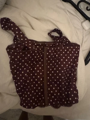 Brun prickig linnetopp - Söt brun linnetopp med vita prickar och breda axelband. Toppen har en lätt croppad passform och dekorativ spetskant längst ner. Perfekt för dig som gillar retrovibbar och vill sticka ut lite extra.