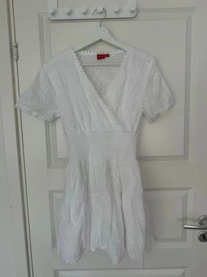 Vit omlottklänning i broderad bomull - Söt vit kortklänning i broderad bomull med omlott och v-ringad hals. Klänningen har korta ärmar, smock i midjan och volangkant nertill. Perfekt för sommarens alla tillfällen och ger en romantisk vibe.💕