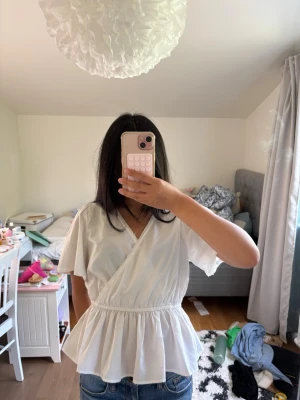 Vit omlottopp från Vero Moda - Säljer en söt vit omlottopp från Vero Moda med korta ärmar och resår i midjan. Toppen har v-ringning och volang nedtill vilket ger en feminin och luftig känsla. Perfekt till jeans eller kjol för en fräsch look.