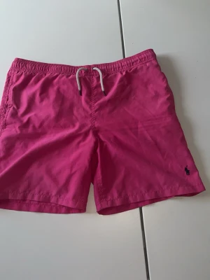 Rosa badbyxor med dragsko - Snygga rosa badbyxor med elastisk midja och vit dragsko. Klassisk modell med kortare ben och liten ficka bak. Materialet är snabbtorkande och perfekt för stranden eller poolen. Liten mörkblå logga broderad framtill.