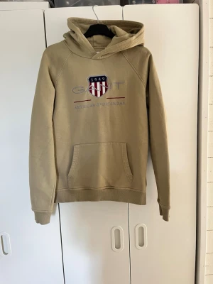 Beige hoodie från GANT med logga 170 - Snygg beige hoodie från GANT med broderad logga och texten 'American Sportswear' på bröstet. Klassisk känguruficka framtill och huva. Mjuk bomullsmix som passar perfekt till jeans eller joggers. Storlek 170