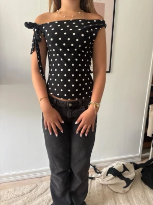 Svart offshoulder topp med prickar - Snygg svart offshoulder topp med vita prickar och knyt på ena axeln. Toppen är tight och har en croppad passform som ger en trendig look. Perfekt för dig som vill sticka ut med ett klassiskt mönster och cool detalj.
