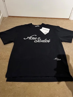 Svart Acne Studios t-shirt med tryck - Snygg svart t-shirt från Acne Studios med vit logga i skrivstil på bröstet och mindre logga nere vid fållen. Klassisk rund hals och korta ärmar. Perfekt till jeans eller shorts för en clean och trendig look.