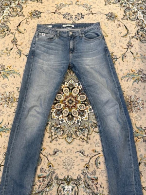 Blå slim fit jeans från Calvin Klein - Snygga blå jeans från Calvin Klein med raka ben och klassisk femficksmodell. Jeansen har en ljus tvätt med subtila slitningar och CK-logga på bakfickan. Tillverkade i mjuk denim som ger en avslappnad look. Storlek 30/32