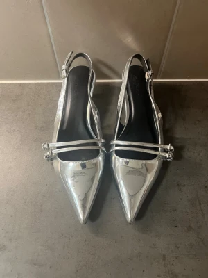 Metallic silver klackskor - Säljer ett par glansiga silvriga pumps med spetsig tå och tunna dubbla remmar över foten. Skorna har öppen häl med justerbar rem och låg klack, 3cm. Materialet är lackliknande och ger en futuristisk vibe. Perfekta för dig som vill sticka ut med metallic på fötterna.